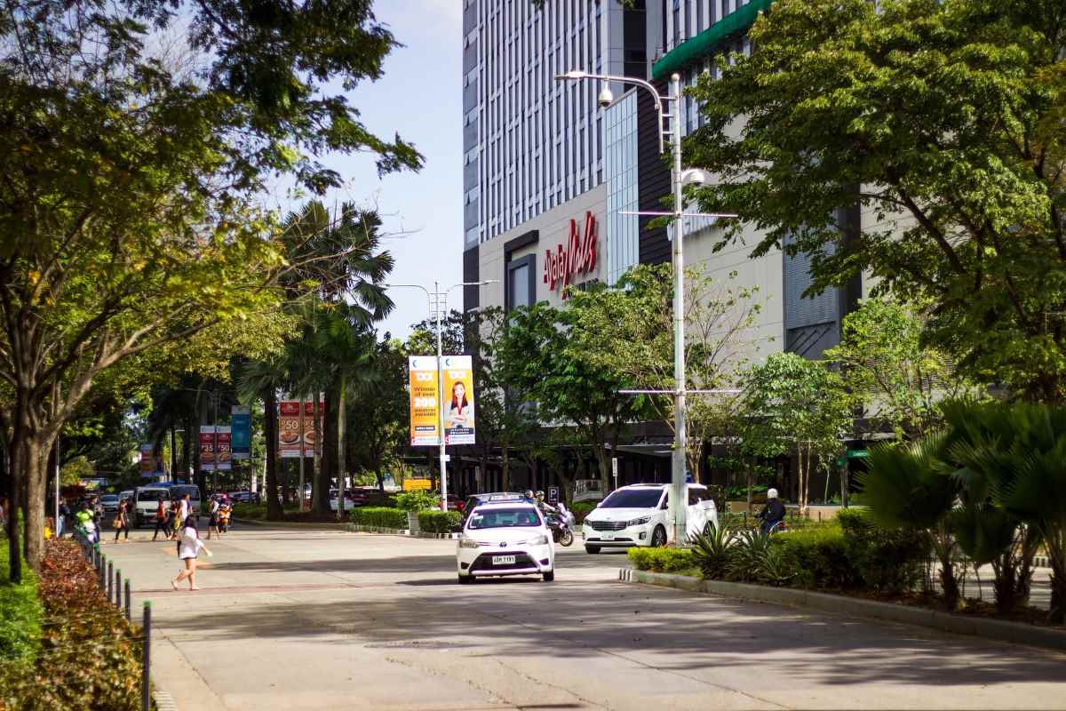 A bright, leafy city street with pedestrians and cars passing a tall glass office building