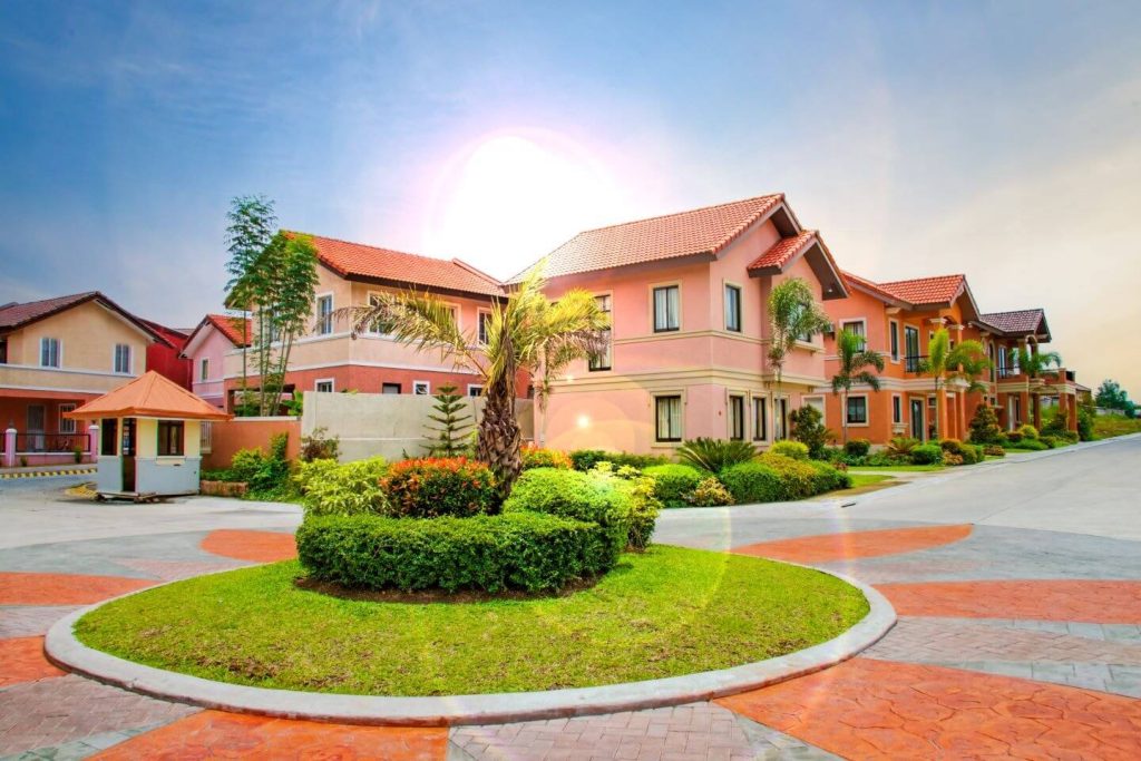 The image shows a well-maintained residential community at Citta Italia by Crown Asia in Bacoor, Cavite, featuring neatly landscaped gardens, modern two-story houses with red-tiled roofs, and clean paved roads under a bright sky, conveying the appeal of this subdivision as a prime real estate properties opportunity in the Bacoor area.
